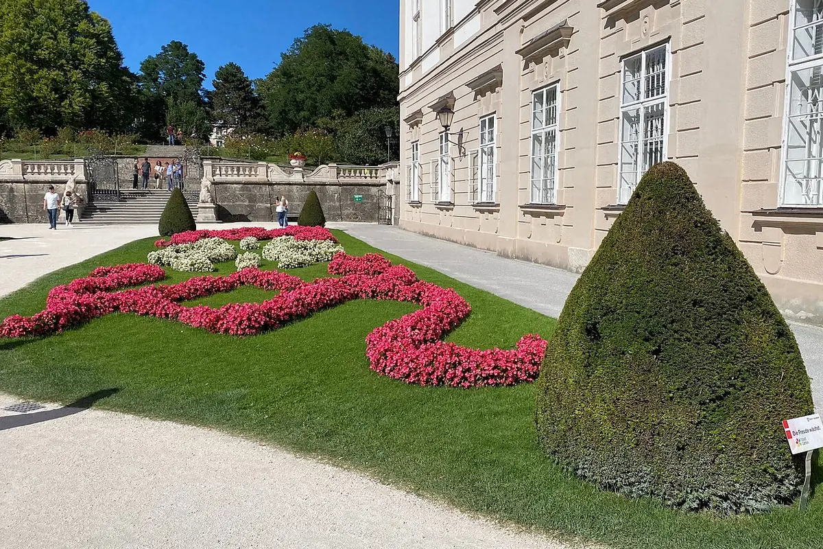 A red and white flower bed and a clipped yew cone beside the palace facade in the Mirabell Gardens, with garden steps beyond