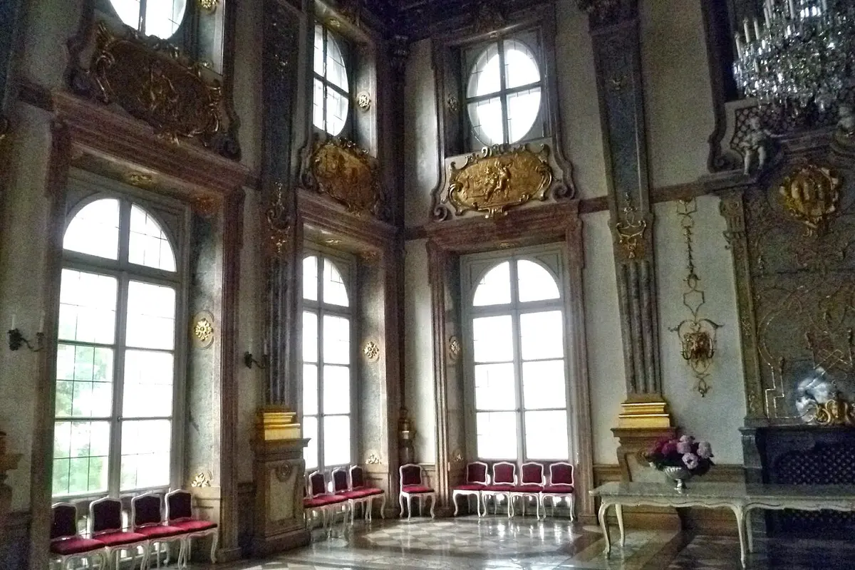 Inside the Marble Hall of Mirabell Palace: tall arched windows, marble pilasters, gilded decoration, a chandelier and rows of red-cushioned chairs on a marble floor